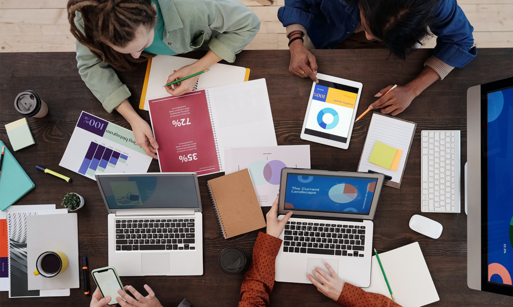 estratégia. Na imagem podemos observar uma mesa com notebooks, tablets e alguns planilhas, nela também podemos ver algumas pessoas como se estivessem em reunião.