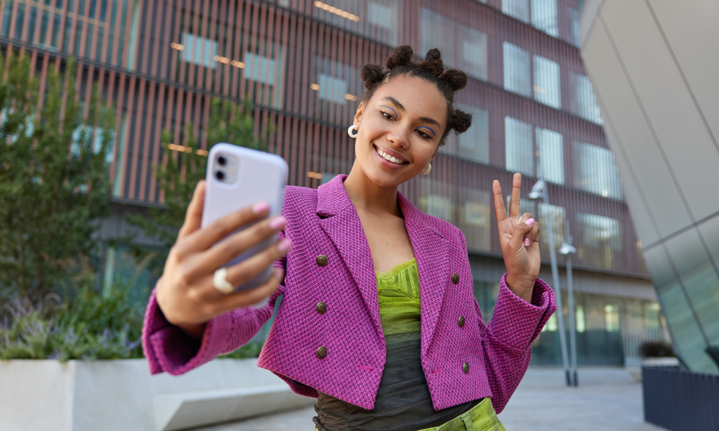 marketing. Imagem de uma menina negra com um vestido verde e blazer roxo fazendo uma selfe na rua. 